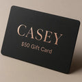 Black gift card with 'CASEY' and '$50 Gift Card' text on a beige background